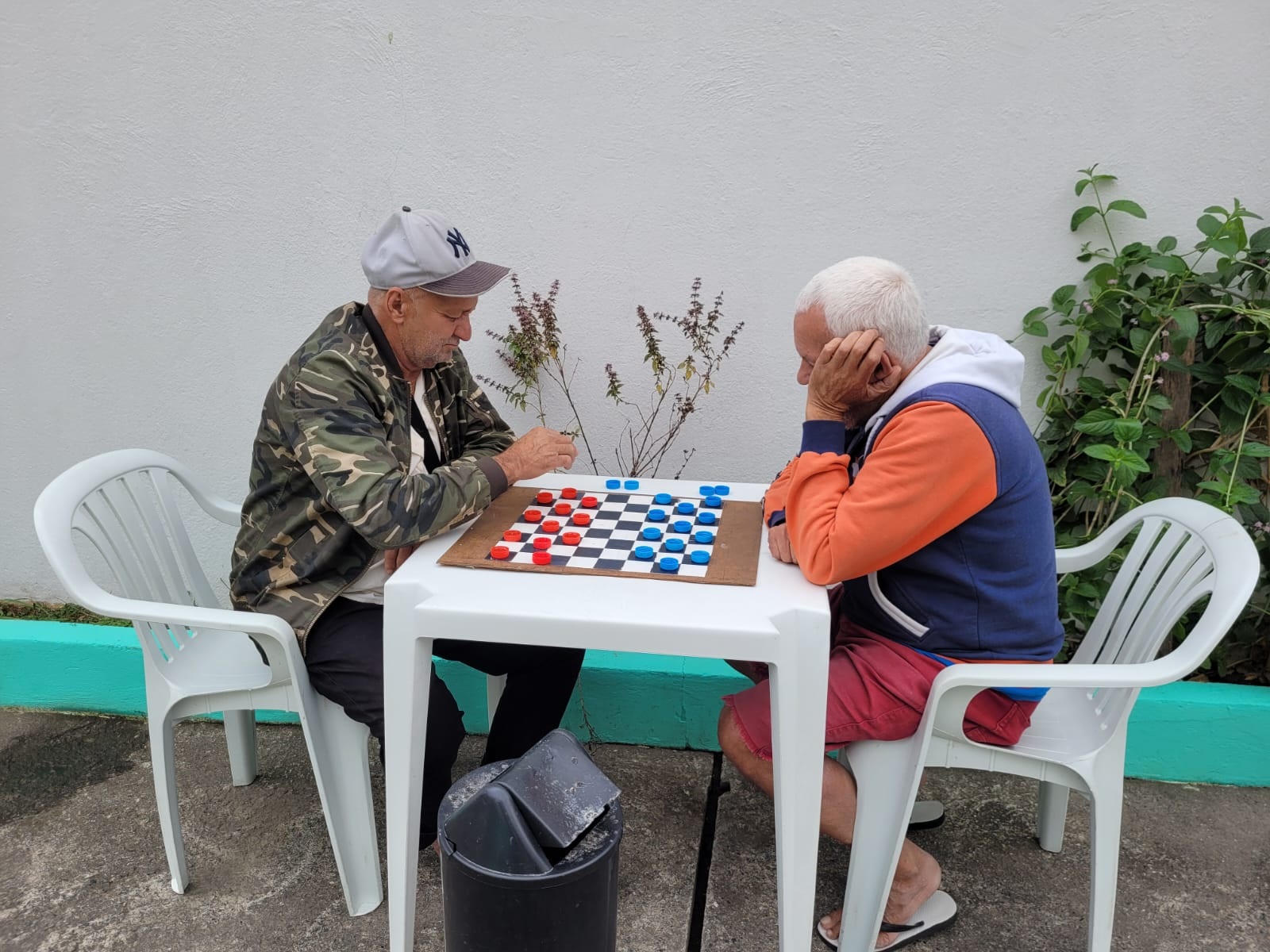 Dois idosos sentados em frente um para o outro jogando uma partida de dama. Tabuleiro em cima de mesa de plástico.