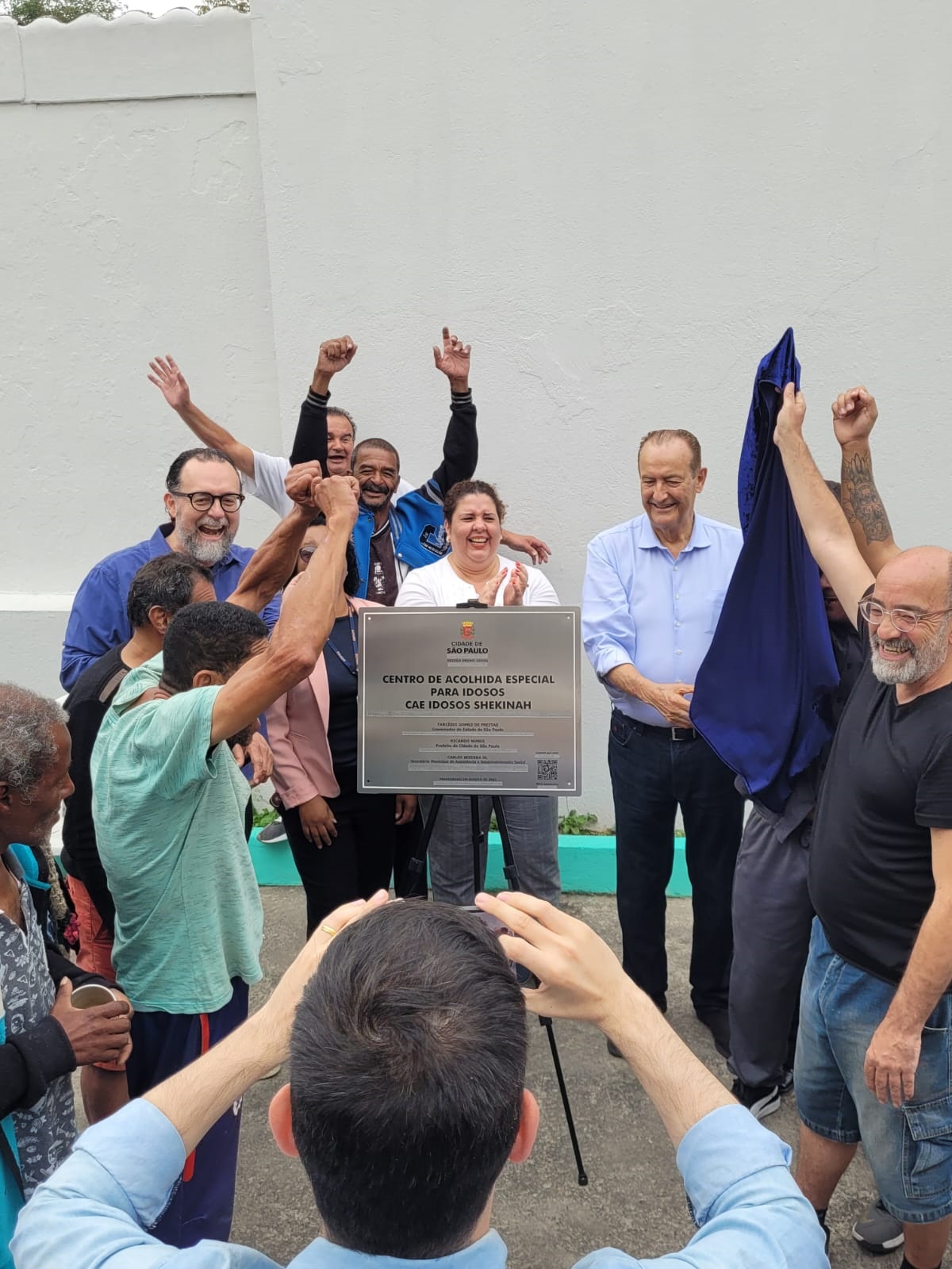 No centro a placa de inauguração do serviço em cima de um cavalete. Ao redor, além do Secretário Carlos Bezerra Jr., outras nove pessoas estão celebrando o descerramento da placa. 
