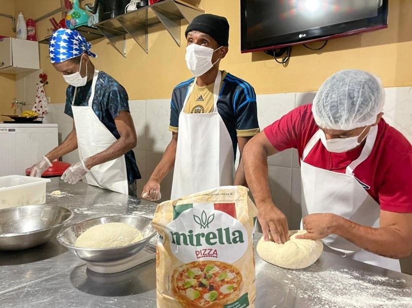 homens de mascara, avental e touca higiênica, manuseando ingredientes durante a aula de gastronomia. Na mesa estão dispostos os ingredientes e utensílios necessários