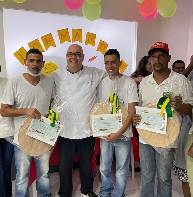 4 homens de camiseta branca lado a lado, 3 deles seguram o certificado de conclusão de curso e junto a eles está o professor Deumas Oliveira