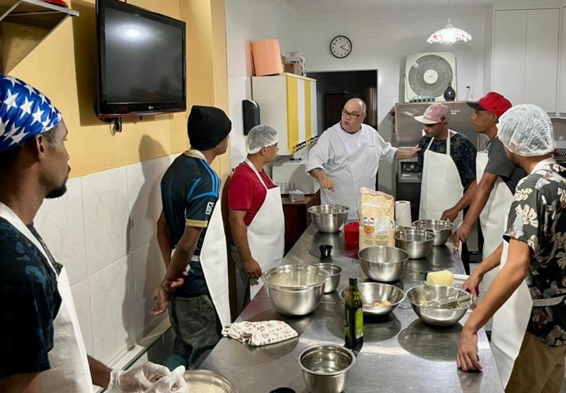 7 homens de avental e touca higiênica durante aula da oficina de gastronomia. Na mesa estão dispostos os ingredientes e utensílios necessários