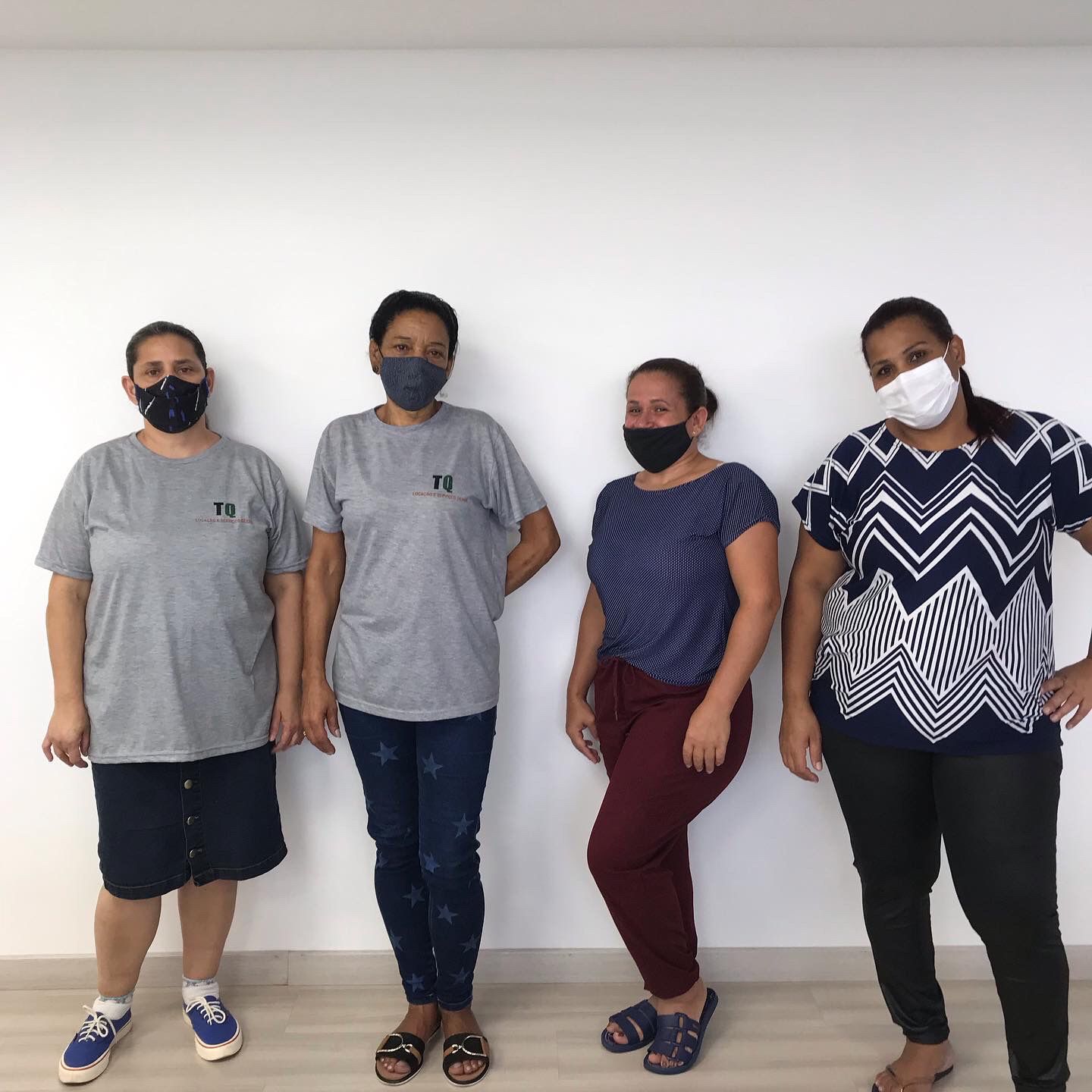 Quatro mulheres em pé, todas com máscara facial. A primeira e a segunda estão com blusas iguais na cor cinza, a terceira veste uma blusa azul marinho e a quarta veste uma blusa preta com listras brancas.