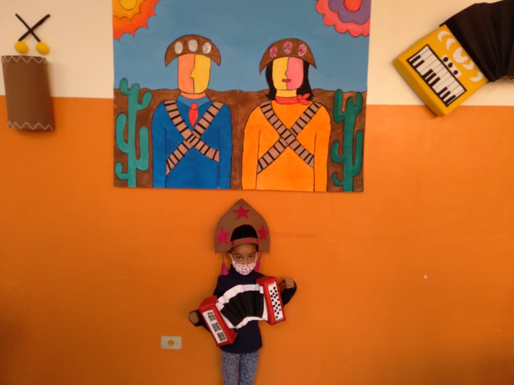 Criança de mascara segurando uma sanfona em frente a uma parede laranja com decorações de temáticas nordestinas