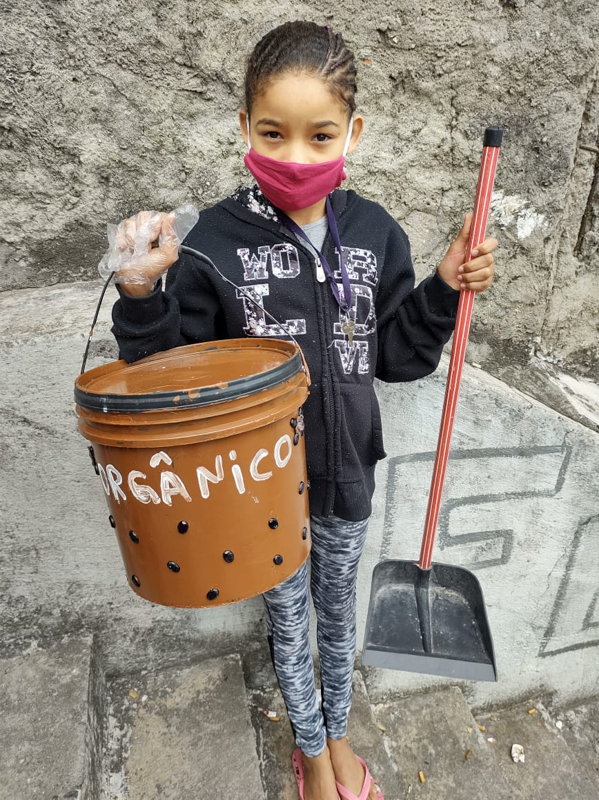 Criança de máscara segurando um balde marrom com a mão esquerda que tem furos na parte de baixo e no centro está escrito orgânico, na mão direita ela segura uma pá. 
