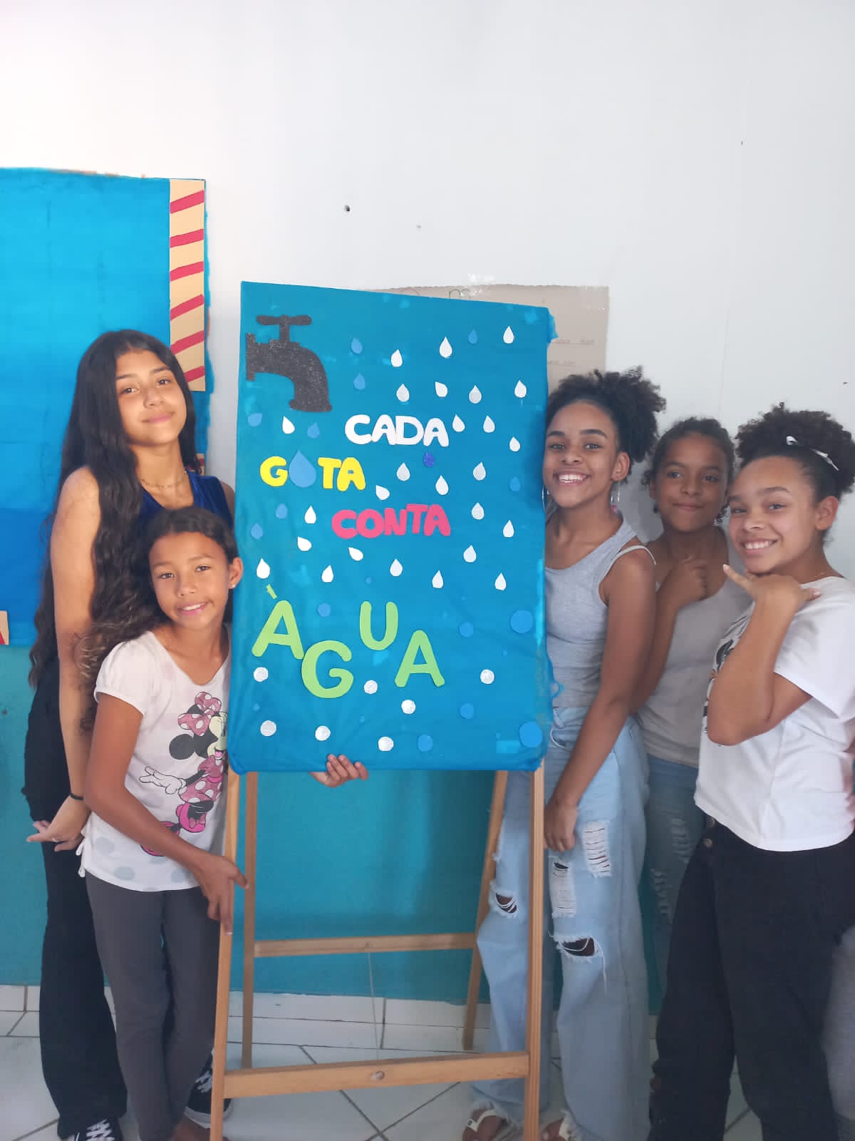 Meninas ao lado de cavalete com um quadro produzido no CCA. Nele há uma representação de uma torneira e gotas d'água. No centro do quadro está escrito 