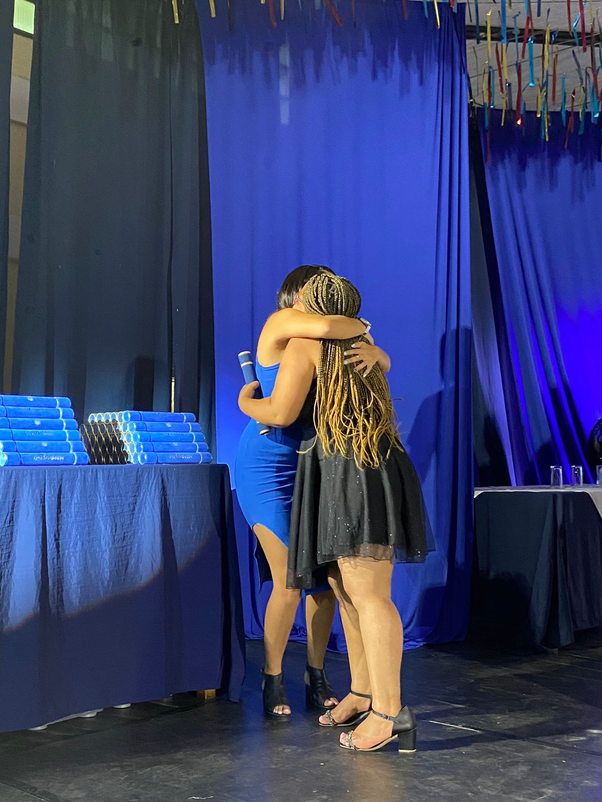imagem de duas mulheres se abraçando. Ao fundo a panos pretos e azuis 