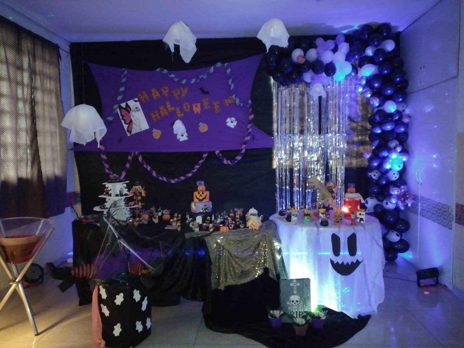 Decoração da festa, ao fundo um painel preto e roxo, na lateral direita tem várias bexigas juntas, no teto tem fantasmas pendurados. Em frente ao painel tem mesas com decorações e doces. 