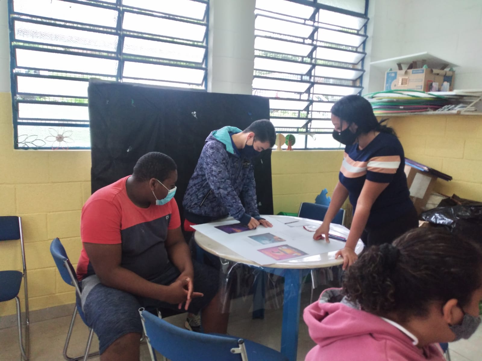 Três jovens de máscara em frente a uma mesa redonda mexendo nos cartazes de decoração. 