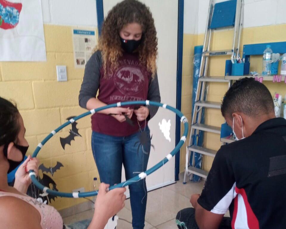 Três jovens de máscara, dois sentados e uma em pé. Eles estão fazendo uma decoração de um bambolê com morcegos e fantasmas pendurados.