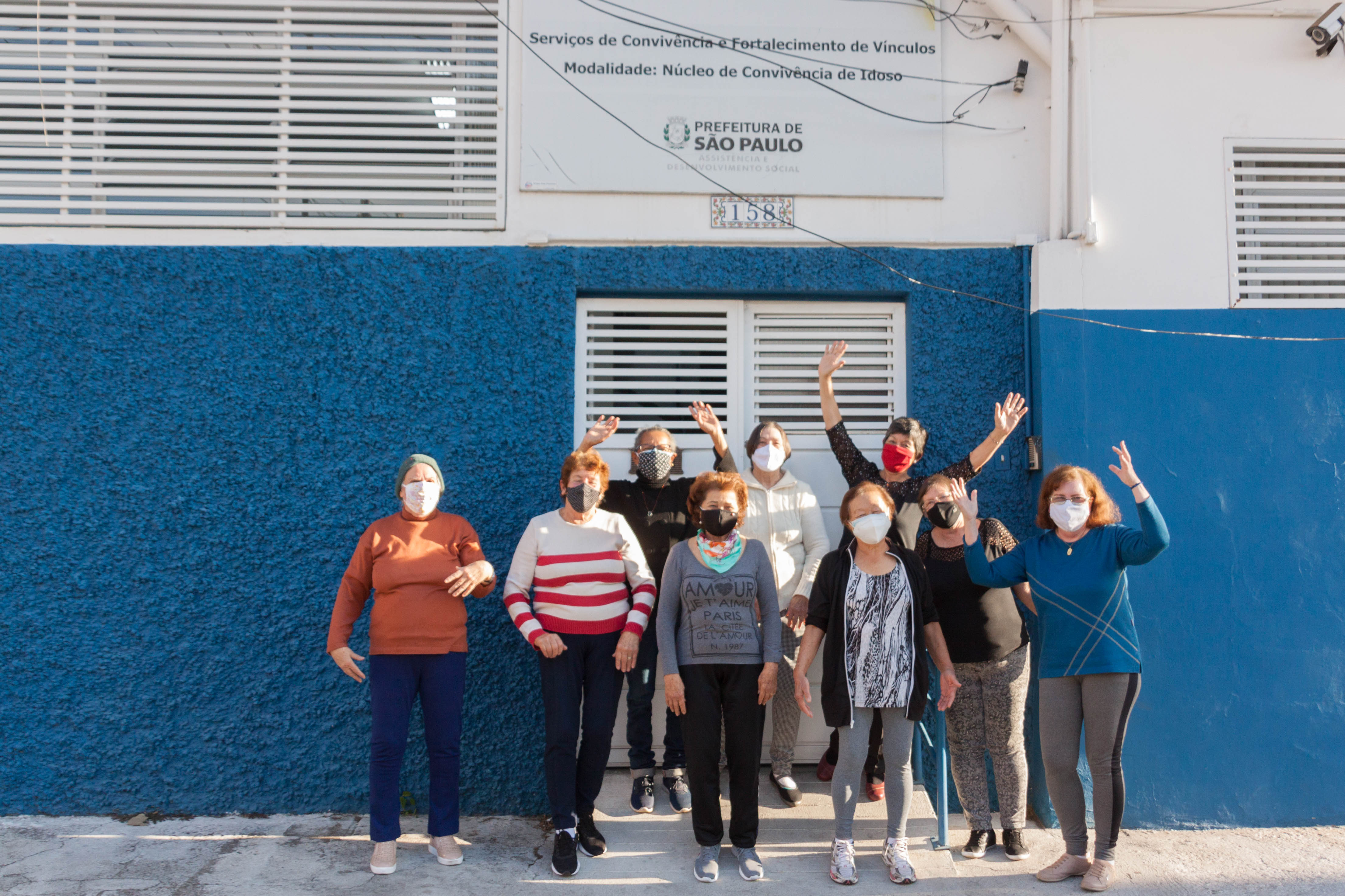 Nove mulheres de máscara em frente a uma parede azul e um portão branco. Três delas estão com as mãos levantadas 