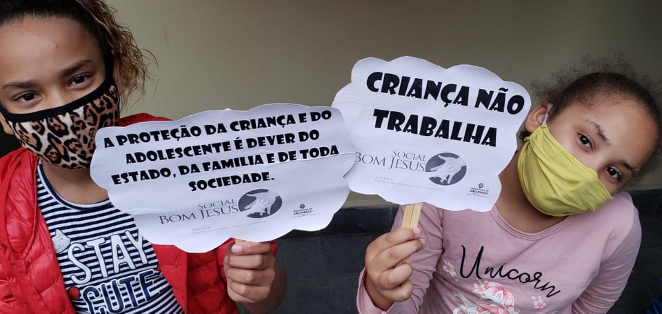 Ao fundo uma parede de tons claros, na frente duas crianças com máscara segurando um cartaz informativo sobre o dia 12 de junho. 