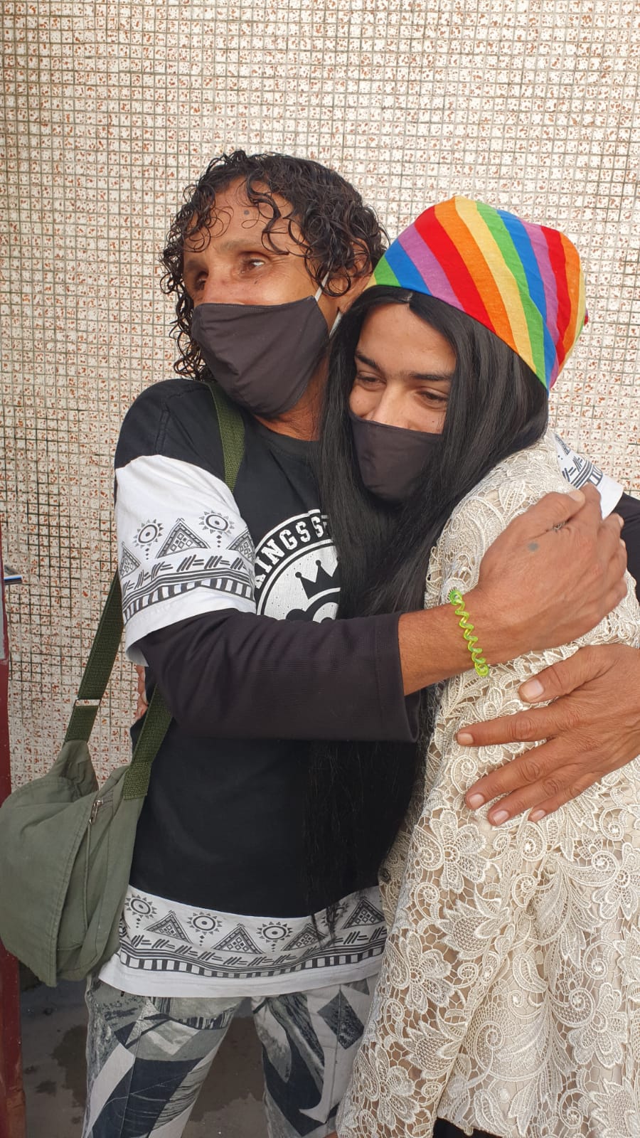 Duas atendidas de máscara abraçadas. Uma delas está com uma bandana com as cores da bandeira LGBTQIA+