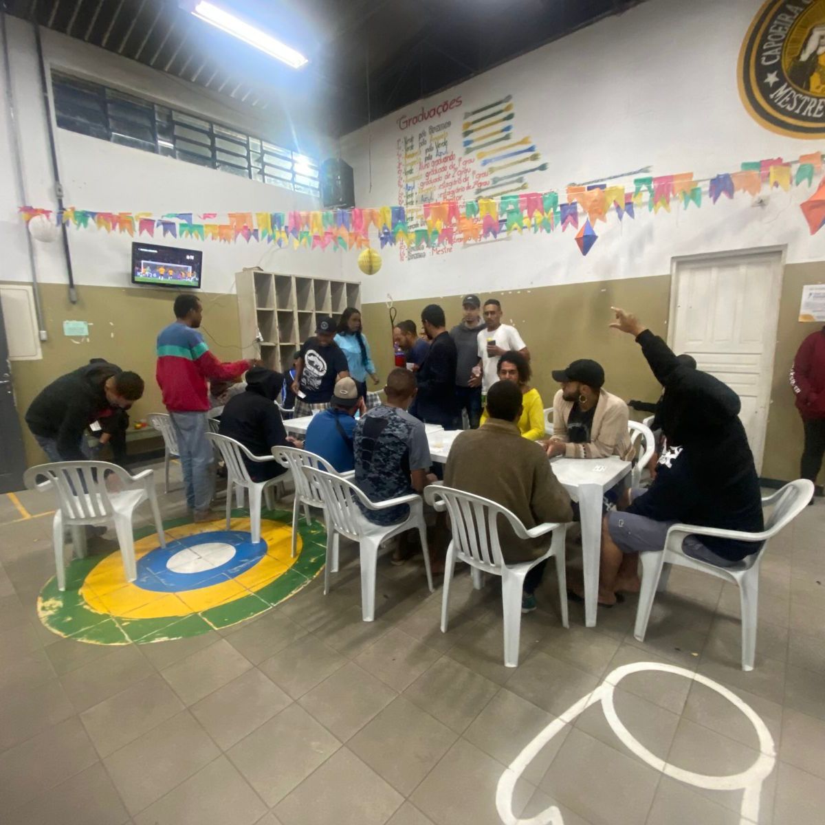 Atendidos durante bingo no serviço. Eles estão reunidos em mesas e cadeiras brancas, e seguram papéis com números. No espaço há bandeiras de festa junina, e as paredes são em tons brancos e marrons.