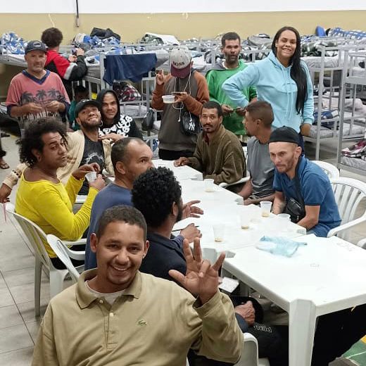 Atendidos sorriem para a foto durante o bingo. Na mesa branca há papéis com números e alguns copos. No fundo há algumas camas com cobertores e travesseiros.