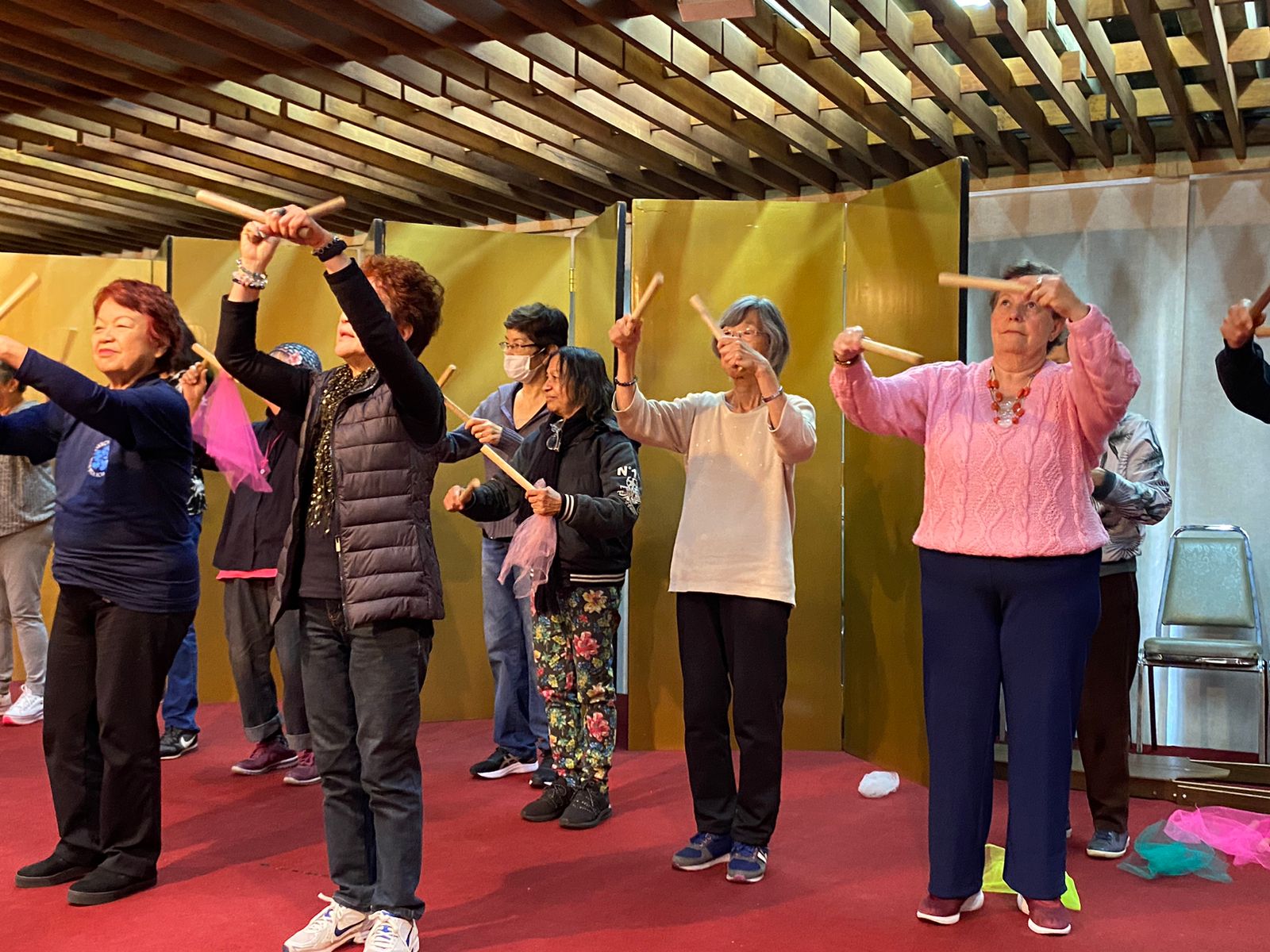 Diversos idosos dispostos lado a lado fazendo a apresentação de uma dança. Cada um deles segura dois pedaços de madeiras, um em casa mão.