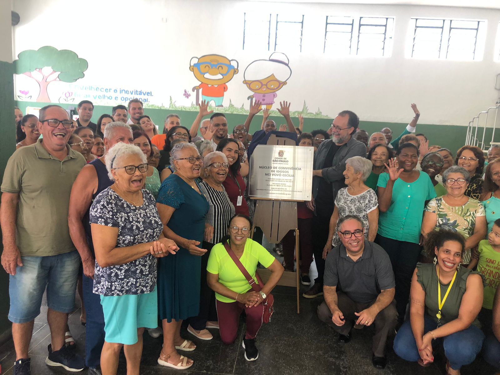 Diversas pessoas envolta da placa de inauguração do novo serviço NCI Vovó Cecília. No fundo haviam perde verde e branca com diversas ilustrações de idosos