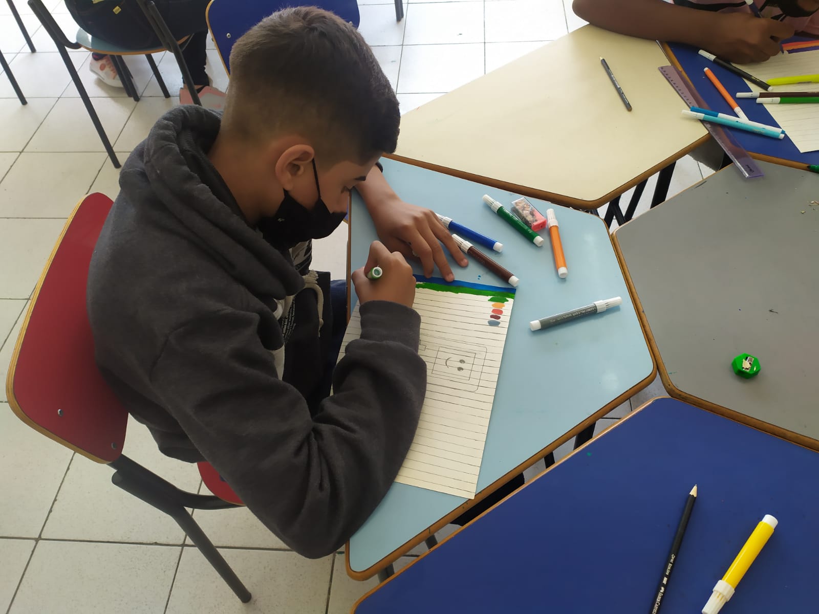 Menino de mascará sentado em frente a uma mesa desenhando em uma folha. Ao redor dele tem lápis e canetas coloridas