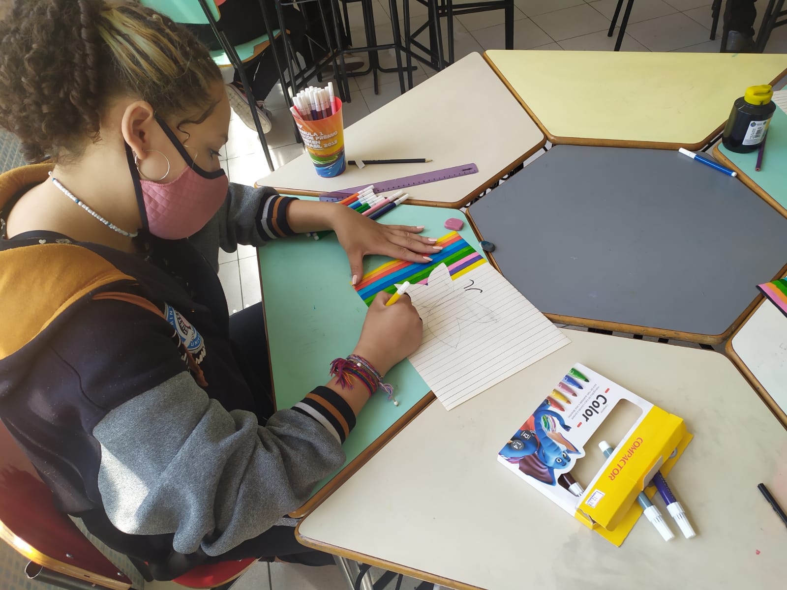Menina de mascará sentada em frente a uma mesa desenhando em uma folha, Ao redor dela tem lápis e canetas coloridas