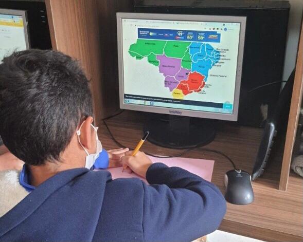 Criança de mascara em frente a um computador com o mapa do Brasil colorido na tela. A criança está com uma caneta em mãos e anotando algo em uma folha de papel rosa