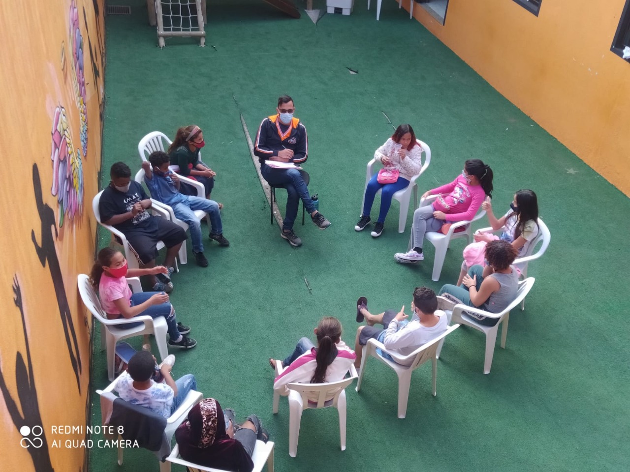13 crianças e adolescentes de máscara sentadas em roda conversando em um ambiente aberto com paredes amarelas e chão verde. 
