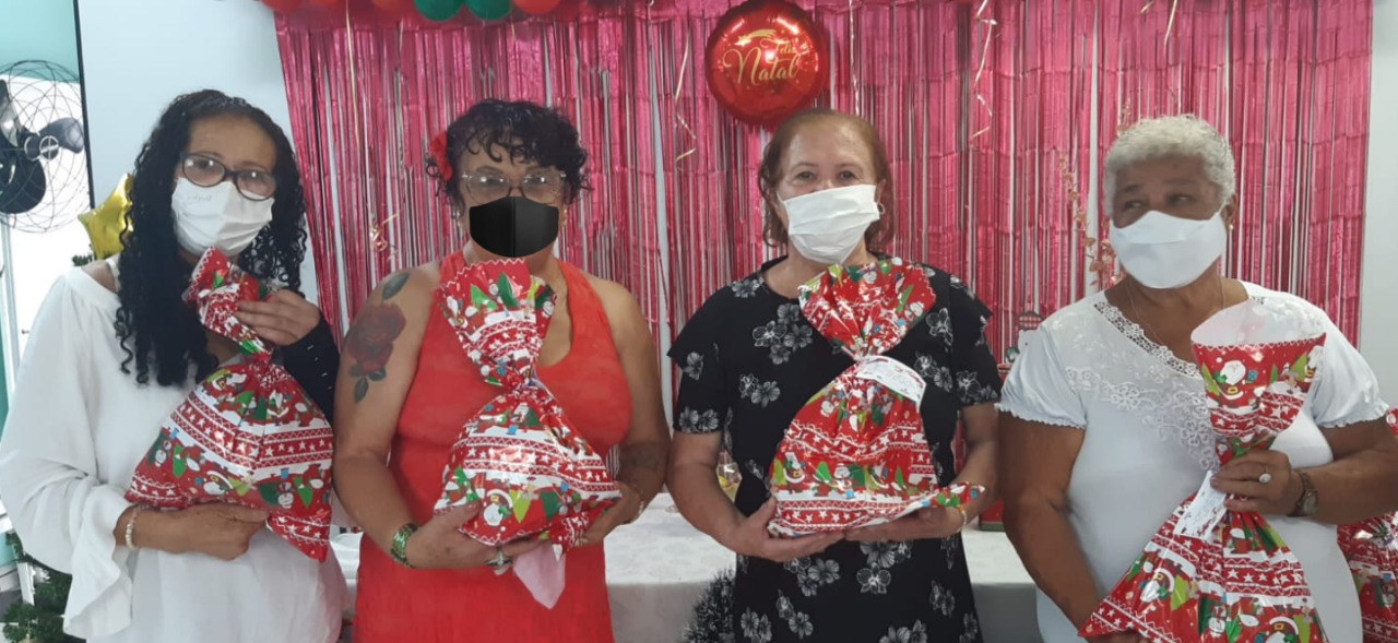 5 senhoras de mascara em frente a uma mesa decorada com artigos natalinos, cada uma delas senhora um pacote de presente 