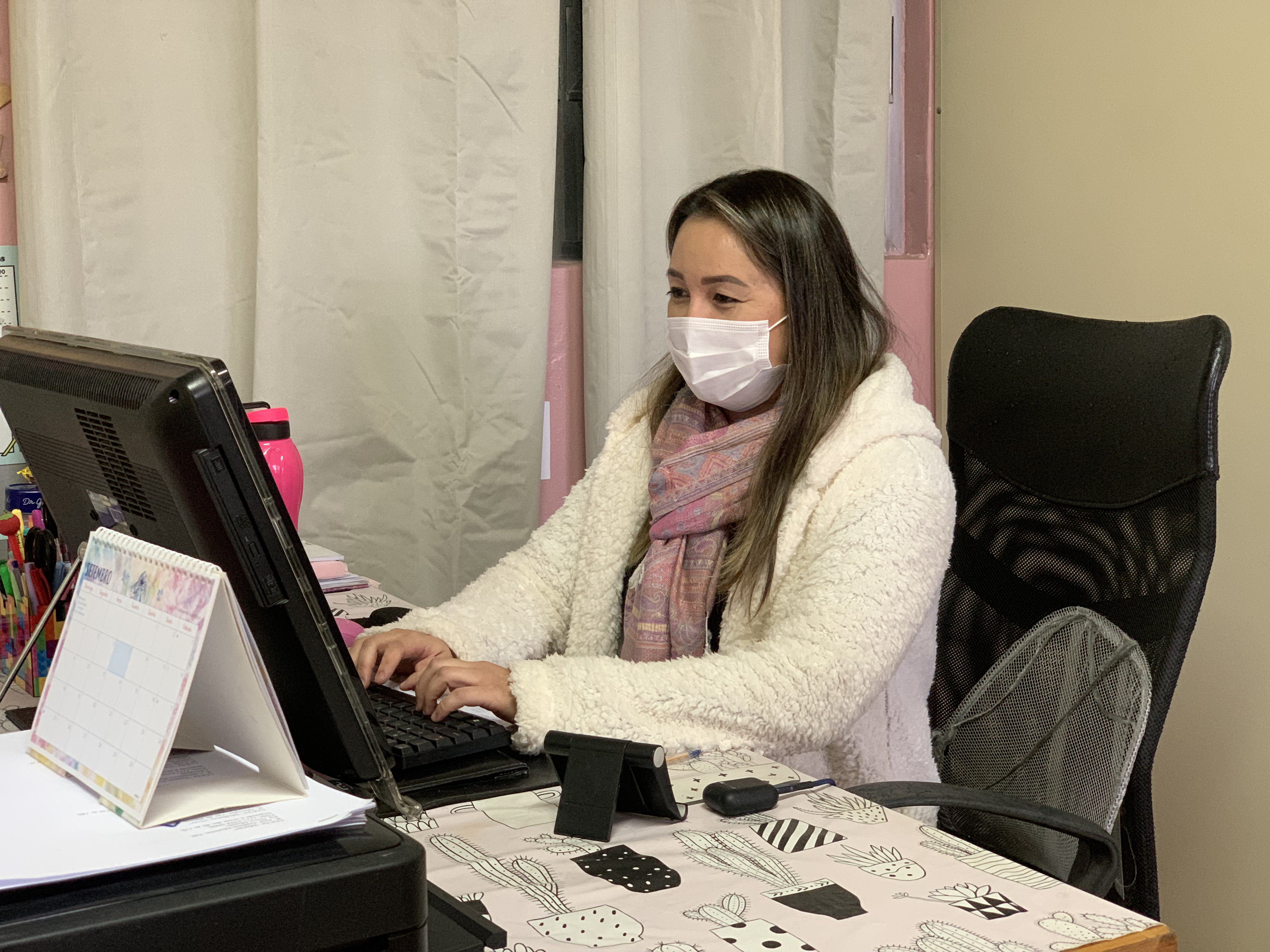 Mulher sentada em uma mesa com com computador olhando-o e digitando. É possível ver em cima da mesa um calendário, um porta canetas e papéis. 