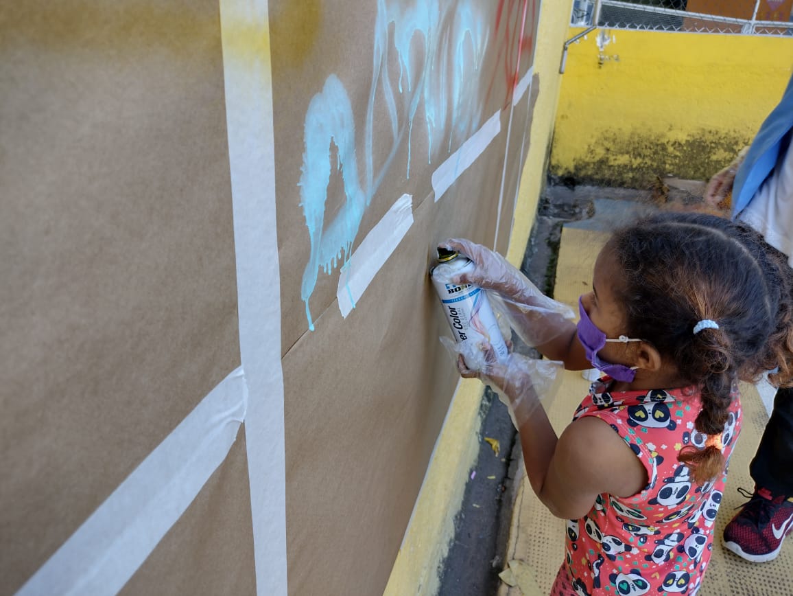 Fundo com parede amarela com uma criança de máscara e luva descartável segurando uma lata de tinta em frente a um mural marrom com desenhos de grafite coloridos. 