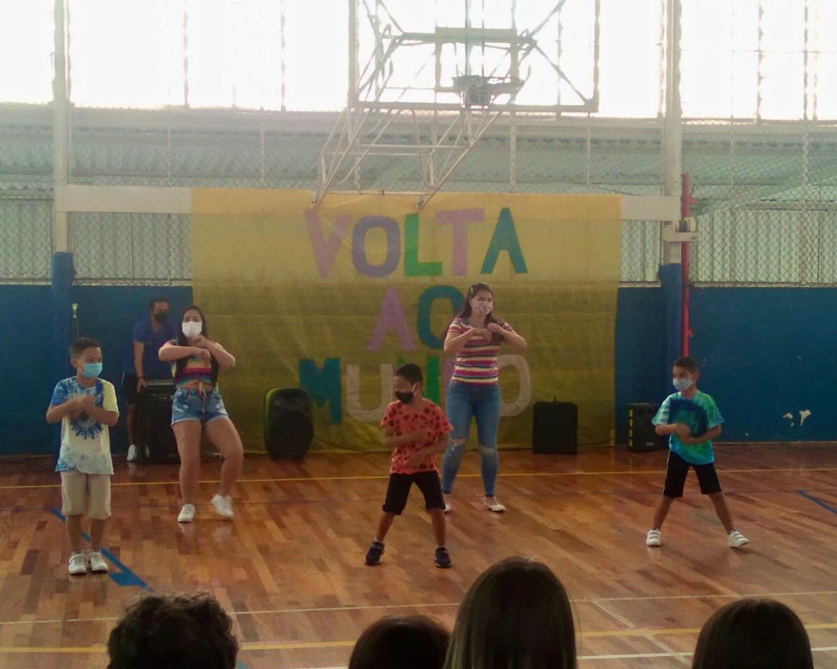 5 crianças de máscara fazendo a sua apresentação. No fundo tem um mural amarelo escrito “Volta ao mundo” em diversas cores 