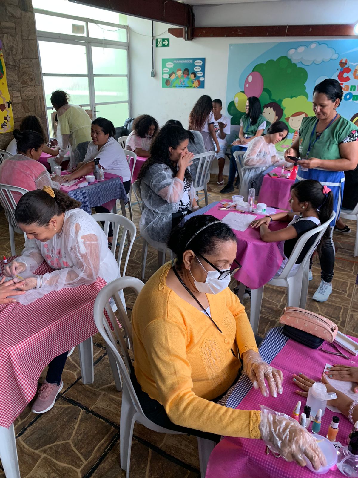 Diversas mulheres estão sentadas em mesas de plástico, cobertas com tecido xadrez e rosa. Elas fazem as unhas, participando da atividade proposta, e conversam. Duas crianças e uma funcionária ao fundo estão de pé conversando. A sala tem em uma das paredes uma decoração com desenhos de crianças brincando. 