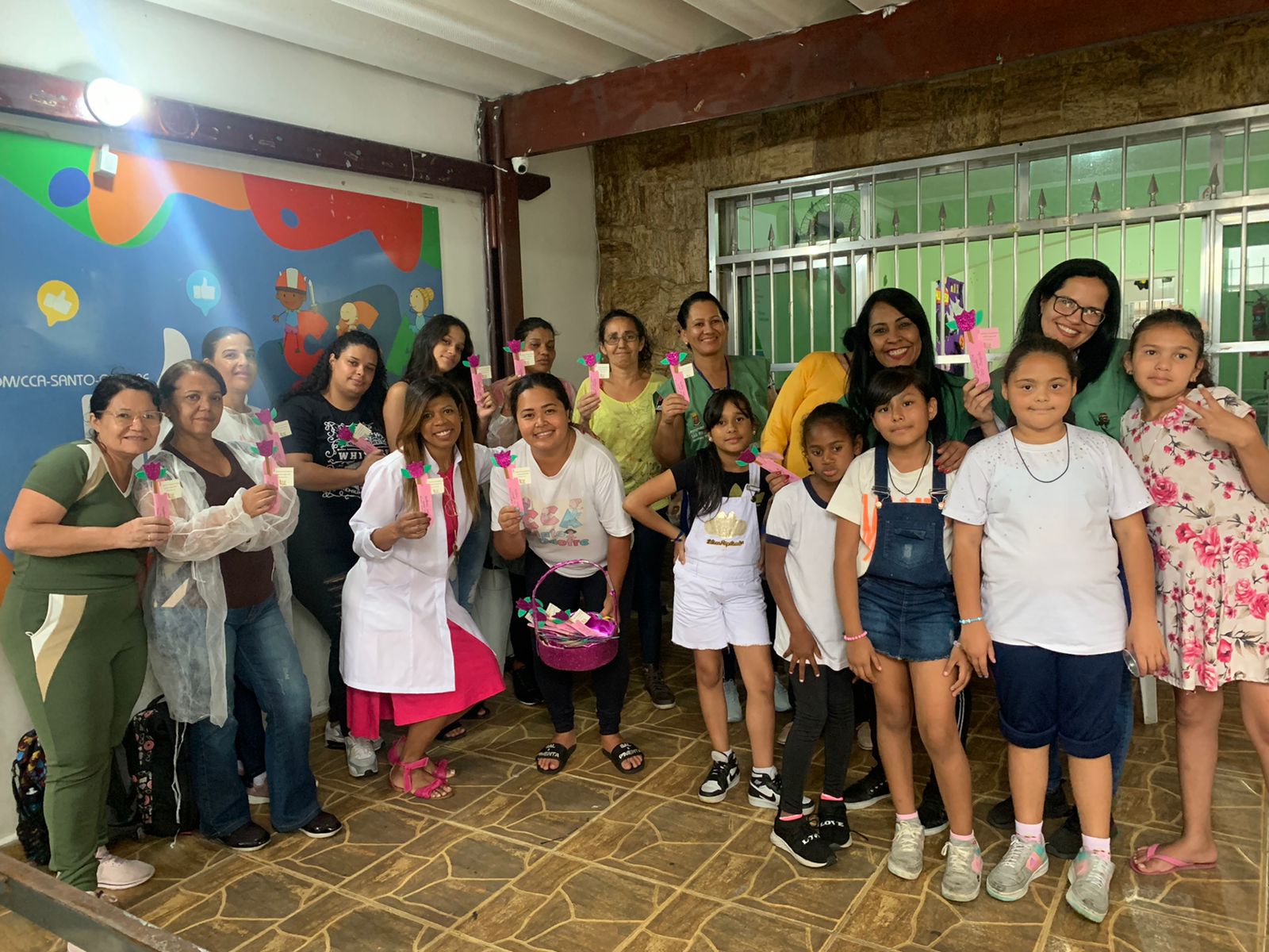 12 mulheres estão de pé ao lado de 5 crianças, todos de pé, segurando as lembrancinhas em suas mãos, confraternizando o final da atividade.