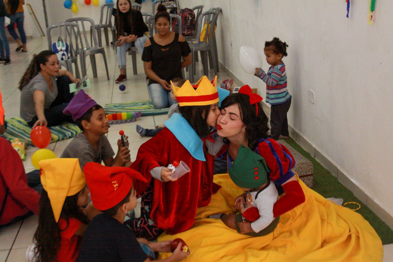 Foto: Crianças sentadas no chão em volta da personagem Branca de Neve e tem criança dando um beijo no rosto da personagem branca de neve. 