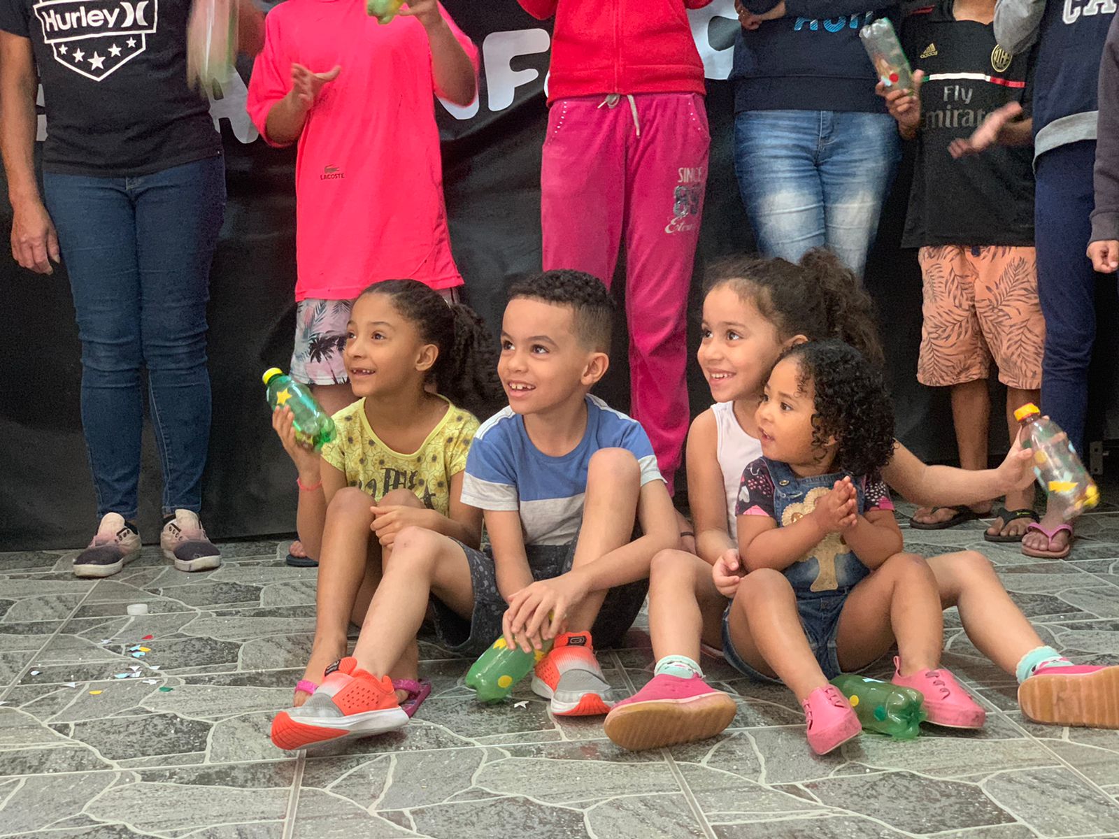 Foto: Quatro crianças sorrindo sentadas no chão lado a lado segurando chocalhos 