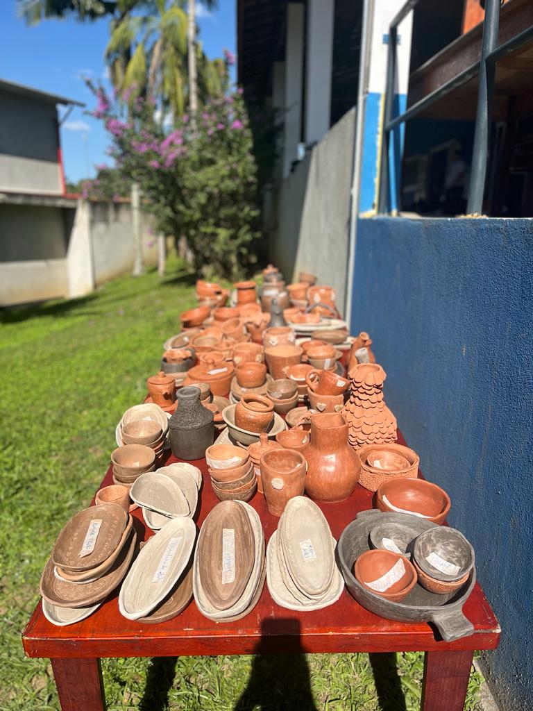 Várias cerâmicas estão dispostas em uma mesa encostada em uma parede. Em destaque estão alguns vasos, pratos, cumbucas e panelas nas cores tradicionais de barro: laranja opaco.