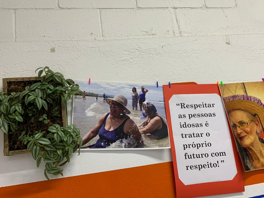 Foto de uma parte da exposição, que mostra uma das acolhidas em uma das viagens desenvolvidas pelo CDI na praia. À esquerda da foto, é possível observar uma planta, está desenvolvida em uma das oficinas de jardinagem pelos próprios idosos; Já, do lado direito, há uma das frases ditas por um dos indivíduos: 
