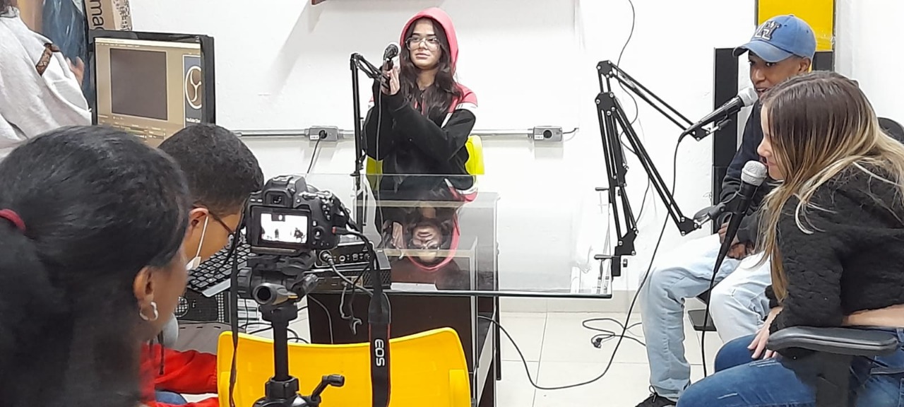 Enquanto são gravados por uma câmera, jovens estão sentados à mesa e produzindo o podcast. Há microfones na frente dos jovens.