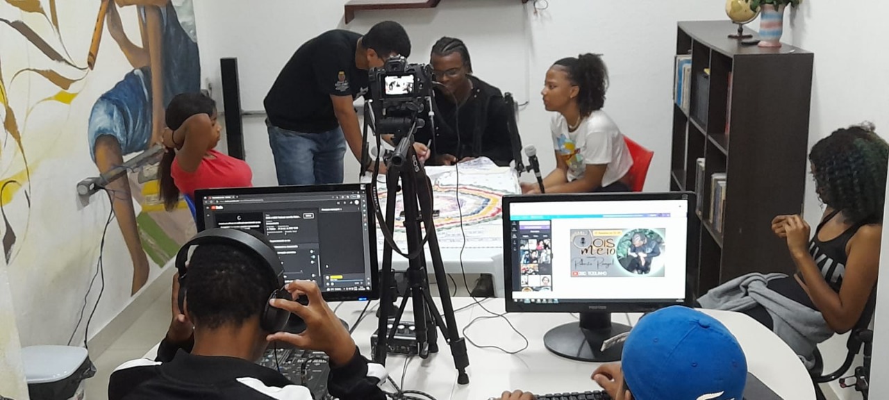 Técnico especializado auxilia convidados e produtores do podcast. Há jovens sentados à uma mesa com microfones, uma câmera e um tripé em sua superfície.