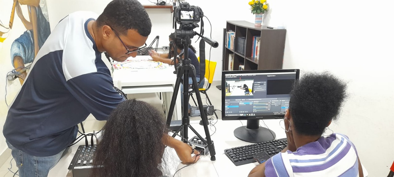Técnico especializado está em pé, auxiliando duas jovens. As meninas estão sentadas à uma mesa, em frente de um computador, uma câmera e um tripé.