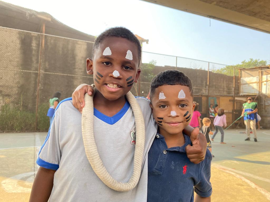 Dois meninos, um vestindo camiseta azul escura e outro branca, estão abraçados, de pé na quadra da comunidade. Eles estão sorrindo para a foto e com a pintura de "coelhinhos" no rosto.