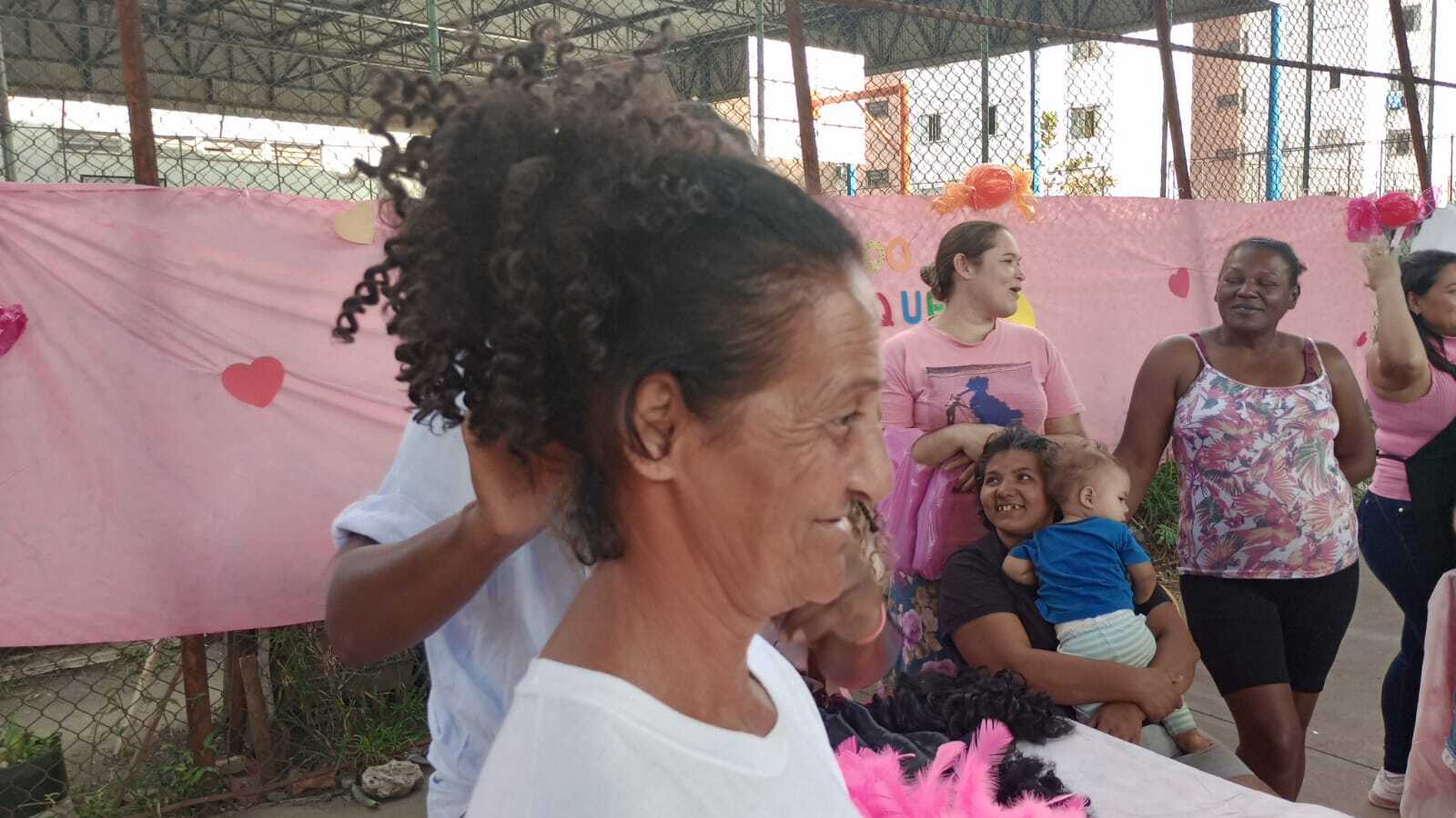 Dona Josefa, está utilizando um aplique de cabelo. Ela está de perfil na imagem, e, outras mulheres estão no fundo observando a oficineira Lígia manipular o aplique no cabelo dela.
