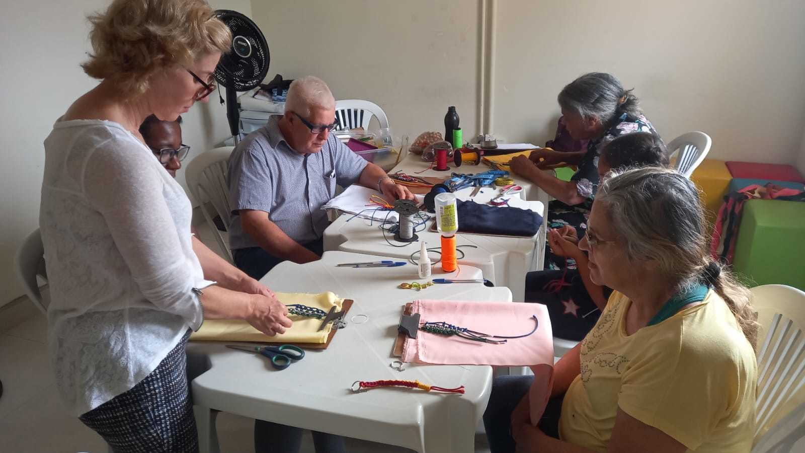 Uma mulher idosa está sentada observando uma outra mulher produzir chaveiros no formato macramê. No fundo, outras duas pessoas produzem outros chaveiros.