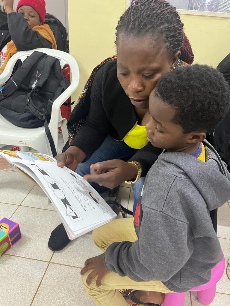 Mulher negra está agachada segurando um livro nas mãos e mostrando para uma criança. No fundo, uma mulher, com touca vermelha, está sentada observando.