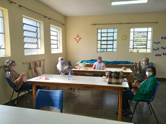 imagem de cinco conviventes durante a aula de artesanato dispostas em duas mesas com máscaras e distanciamento social. 