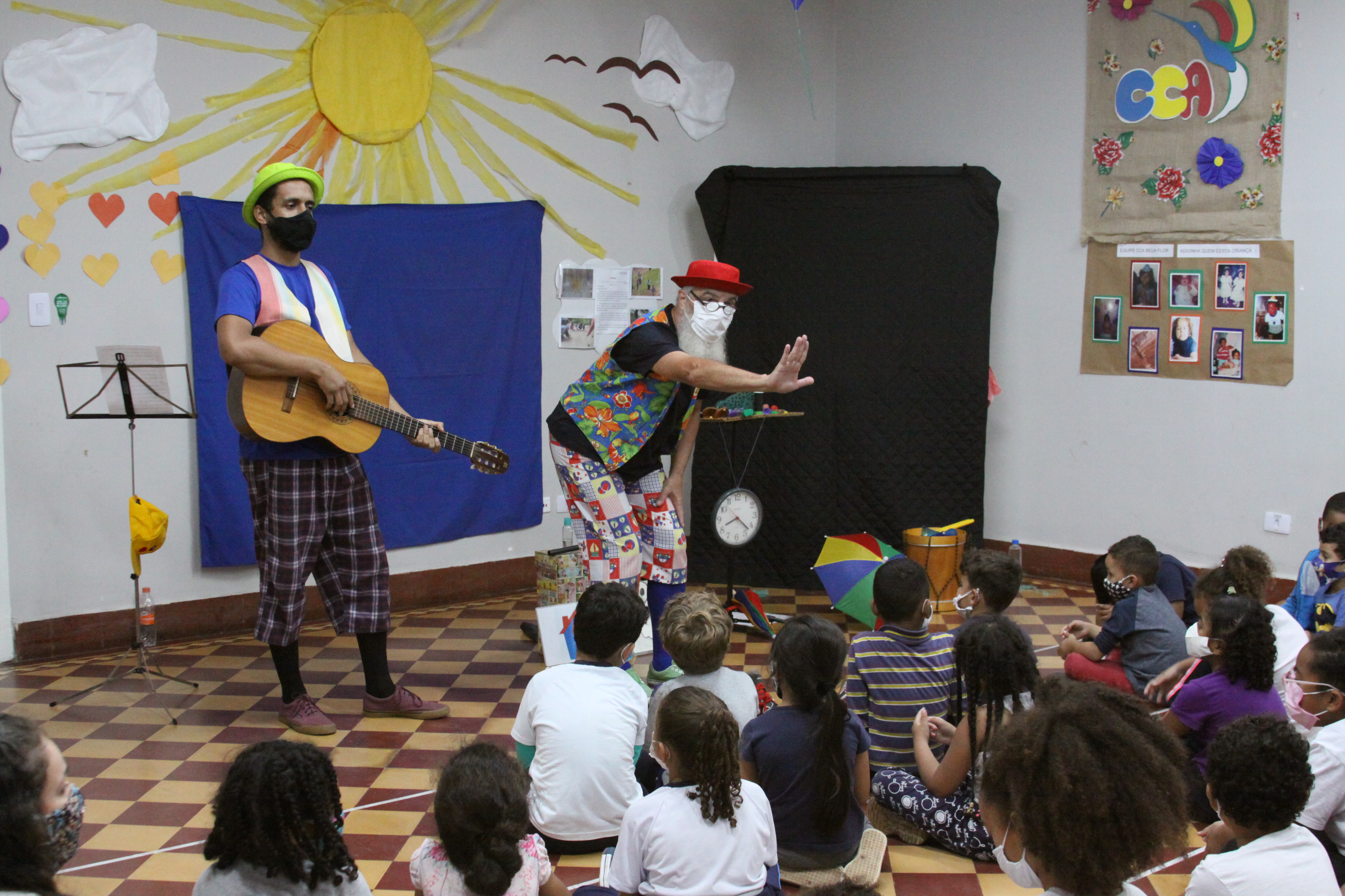 Na parte inferior da imagem estão as crianças sentadas no chão de costas para a câmera e em frente a eles estão os dois artistas de pé, o da esquerda está tocando violão.
