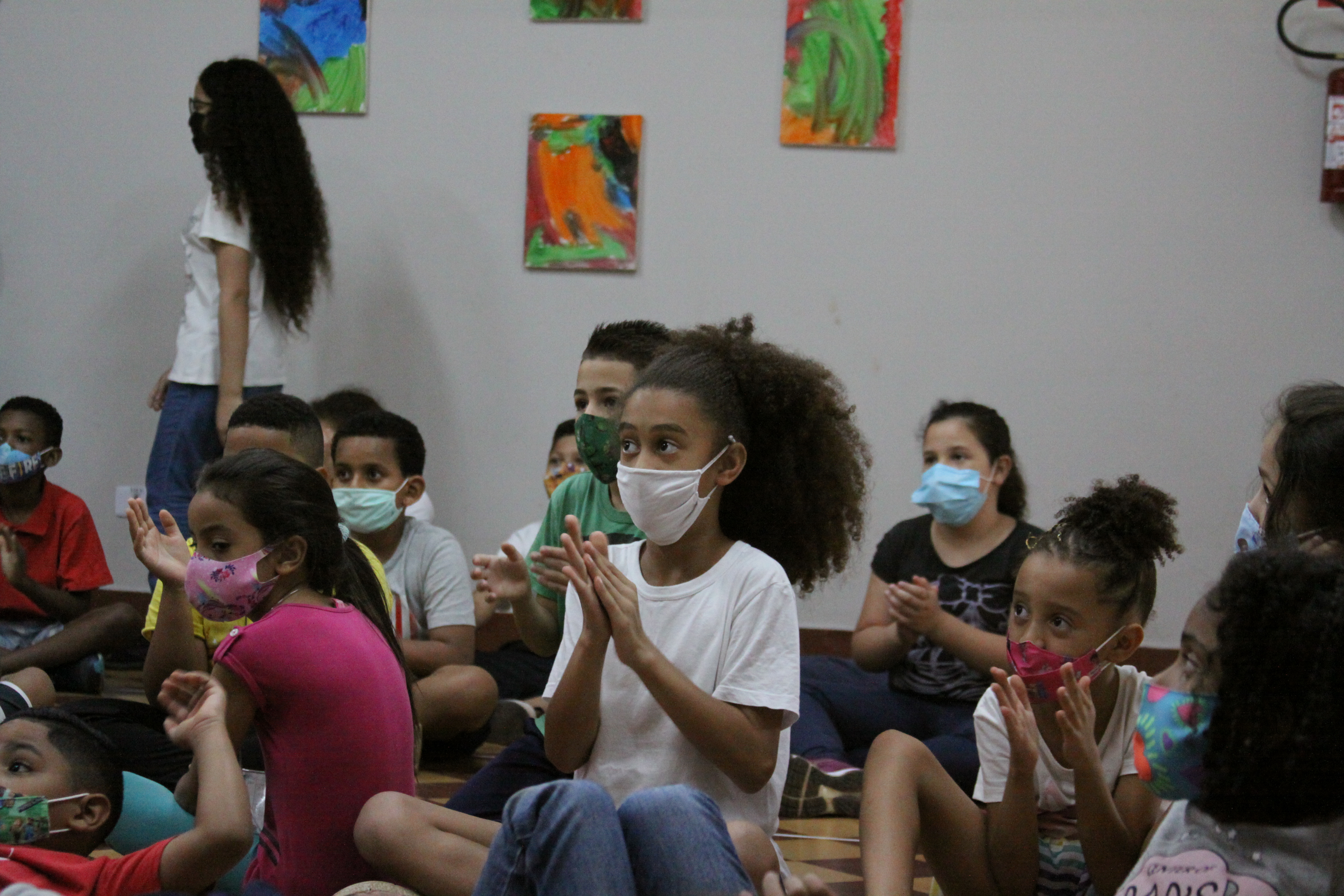 Imagem de cerca de 15 crianças sentadas assistindo à apresentação, no centro da foto há uma menina batendo palma.