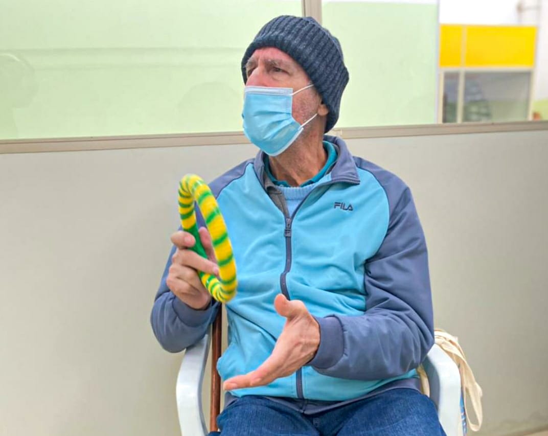 No centro da imagem está um idoso atendido pelo CDI com um instrumento circular em sua mão. Ele está sentado em uma cadeira branca e olha para o lado esquerdo da foto. 