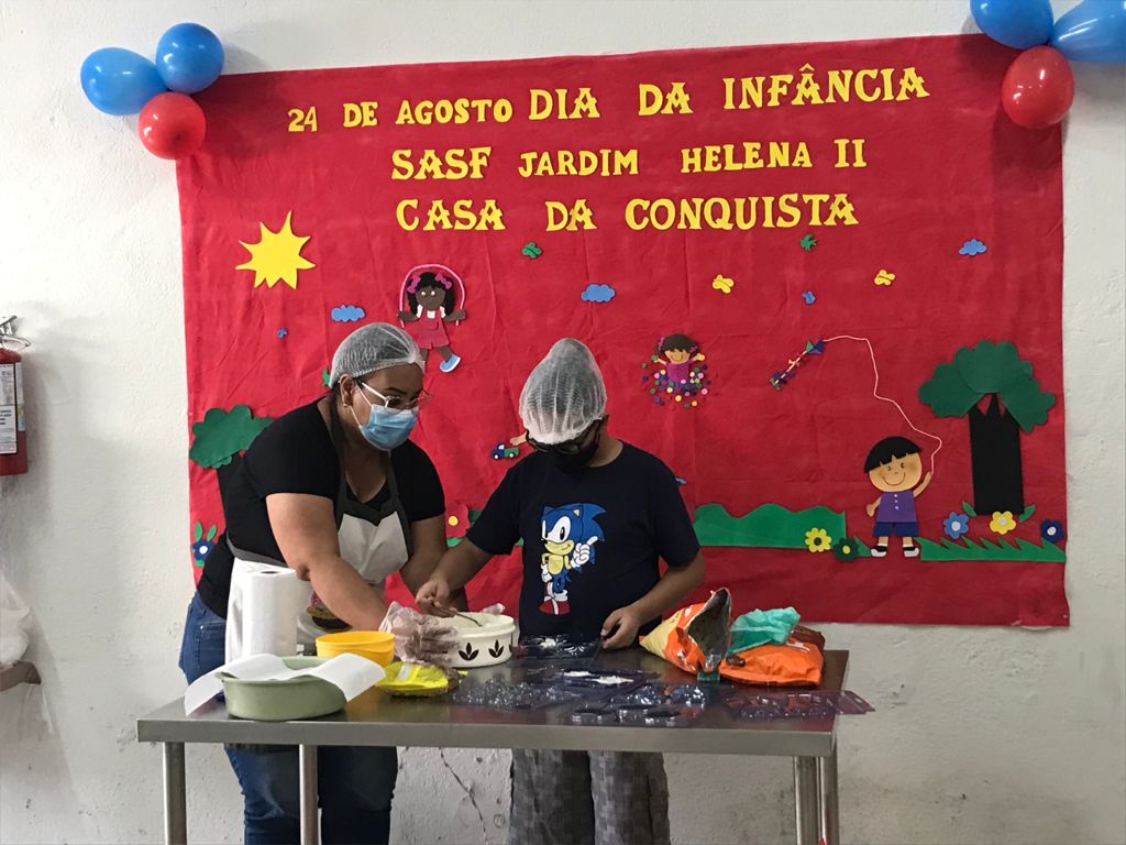 Foto de uma mulher e um menino atrás de uma mesa enquanto ele despeja o chocolate nas forminhas para fazer os pirulitos. Na parede ao fundo está um painel com o título “24 de agosto - Dia da Infância SASF Jardim Helena II Casa da Conquista” e colagens de E.V.A. com desenhos de crianças brincando.