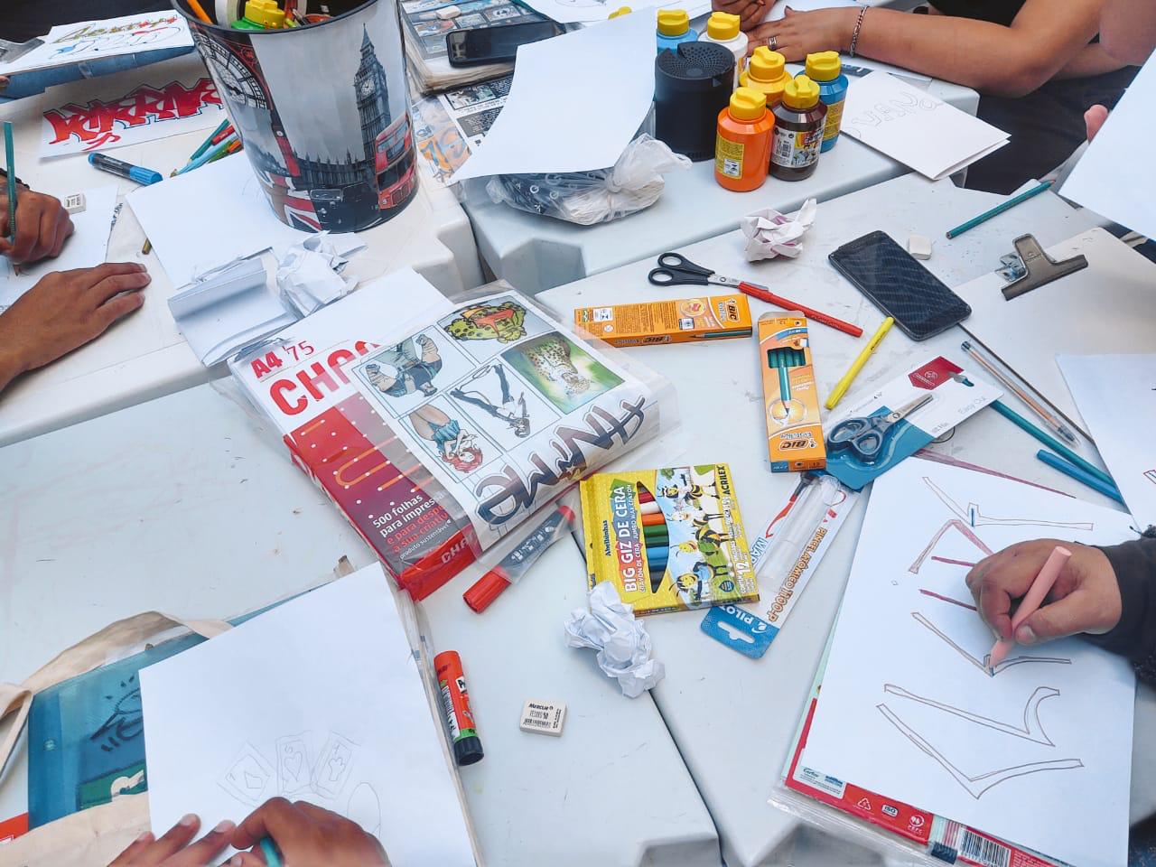 foto da mesa com materiais dispostos enquanto as mãos de quatro jovens aparecem desenhando nos quatro cantos da imagem