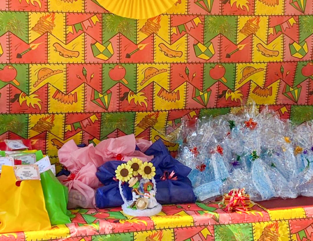 Foto da mesa dentro da tenda com toalha decorada com bandeirinhas juninas e com diversos kits colocados em cima dela, no centro da foto também está um enfeite com girassóis artificiais