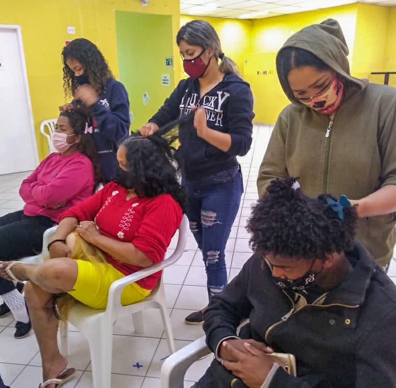 Imagem de três atendidos sentados lado a lado enquanto têm seus cabelos trançados por outros três participantes da oficina.