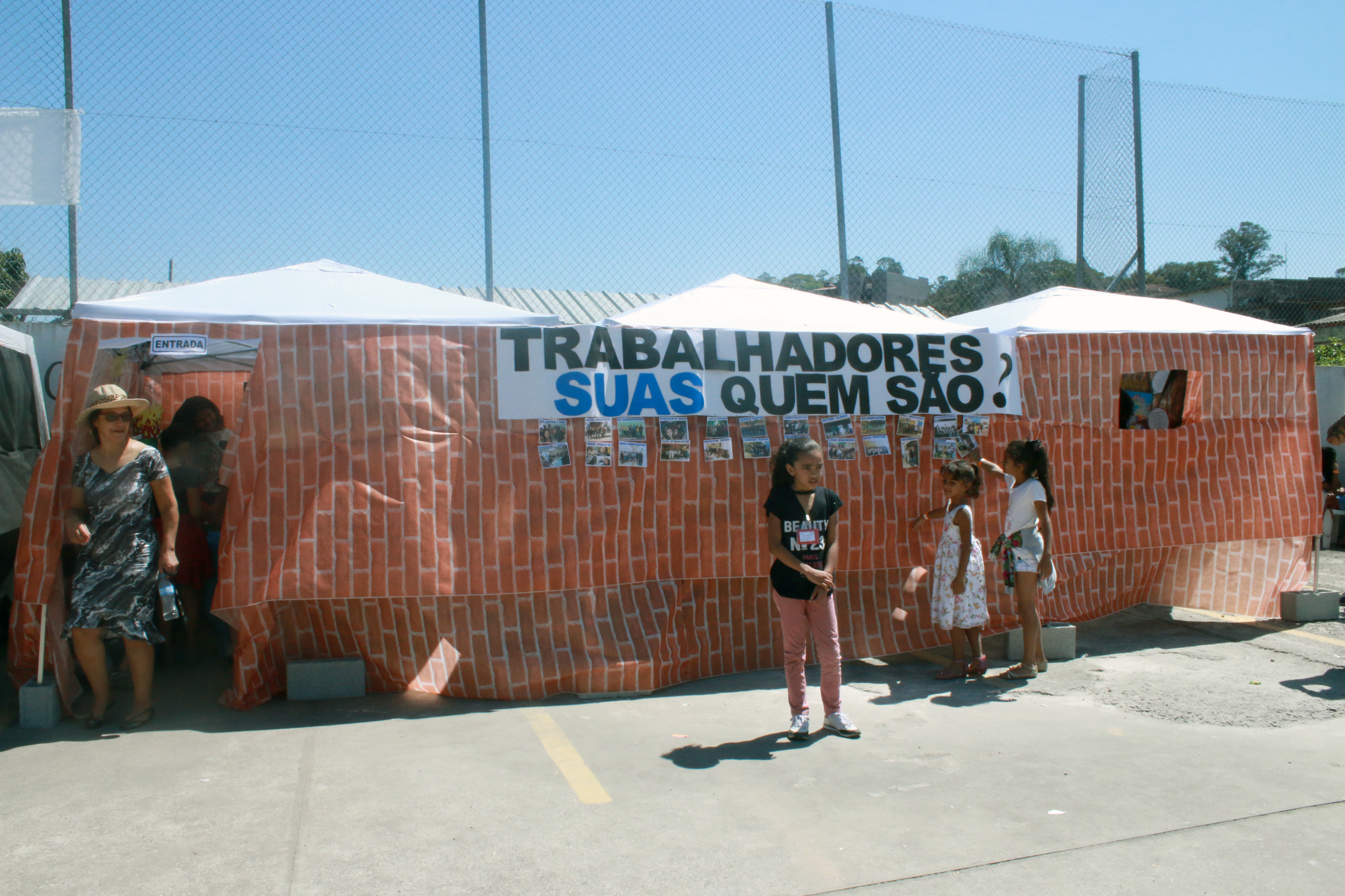 Três crianças estão na frente de três tendas que estão cobertas por uma lona com desenho de tijolos. Na lona há uma faixa com a frase “Trabalhadores SUAS Quem São?” com imagens coladas em baixo.